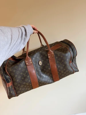 Weekendbag  - Säljer en stor och snygg weekendbag med klassiskt monogrammönster i mörkbrunt och beige. Väskan har detaljer och handtag i brunt skinn, dragkedja och en praktisk namntag. Perfekt för weekendresor och har riktigt lyxig vibe. Har en del effekter. Finns fler bilder vid intresse 
