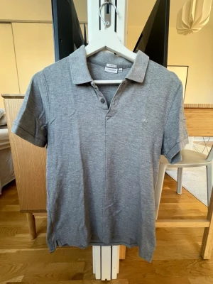 Grå piké från J.Lindeberg slim fit - Säljer en stilren grå pikétröja från J.Lindeberg i slim fit-modell. Tröjan har klassisk krage, korta ärmar och knappar vid halsen. Perfekt för dig som gillar enkel och snygg design med diskret logga på bröstet.