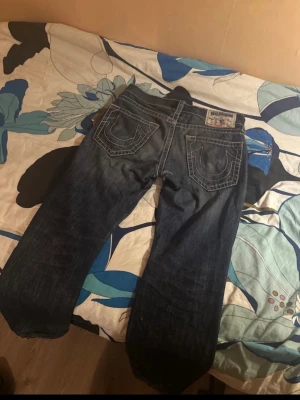 True Religion blå jeans, storlek 31 - Säljer ett par blå jeans från True Religion med ikoniska bakfickor och kontrastsömmar. Modellen har rak passform och klassisk femficksdesign. Jeansen är tillverkade i denim och har detaljer som slitningar och grova sömmar. Perfekt för dig som gillar streetwear och vill sticka ut.