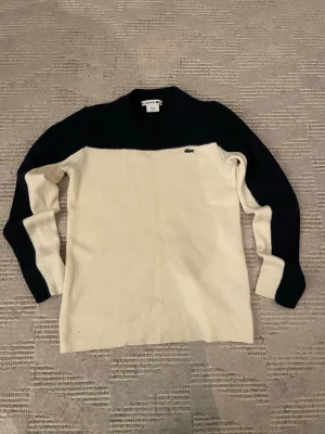 Vit och marinblå stickad tröja Lacoste - Snygg stickad tröja från Lacoste i vit och marinblå färg. Klassisk design med rund hals och långärmade ärmar. Tröjan har det ikoniska Lacoste-krokodilemblem broderat på bröstet. Perfekt för dig som gillar stilrena och sportiga plagg.