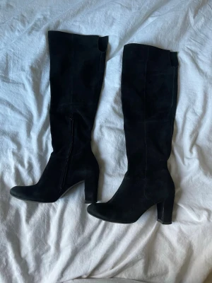 Svarta höga mockastövlar med klack - Säljer ett par svarta stövlar i mocka med rund tå och hög klack. Stövlarna har dragkedja på insidan för enkel på- och avtagning. Stilrena och passar perfekt till höst och vinter. Snyggt material och klassisk form.