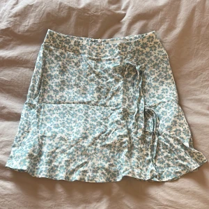 Blommig omlottkjol från Pull&Bear - Enen söt kortkjol från Pull&Bear i storlek S. Kjolen är vit med ljusblått blommönster, har volangkant och omlott med knytband i sidan. Materialet är mjuk och lätt viskos, sval och inte alls genomskinlig. Saknar helt slitage och är använd väldigt sparsamt.