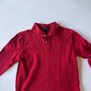 Röd långärmad polotopp från Polo Ralph Lauren - Säljer en klassisk röd långärmad polotopp från Polo Ralph Lauren med krage och två knappar framtill. Toppen har en broderad blå logga på bröstet och är tillverkad i mjuk bomull. Perfekt för en stilren och sportig look.