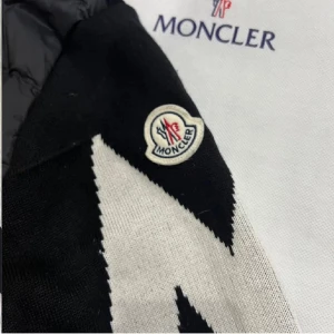 Svart Moncler cardigan  - Säljer en svart pufferjacka från Moncler med huva och snörning. Jackan har en dragkedja framtill och en unik vit zigzag-detalj på ena ärmen samt Moncler-logga. Materialet är en mix av polyester och stickad textil, perfekt för dig som vill ha en snygg och varm jacka.