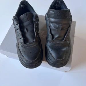 Svarta Nike Air Force 1 sneakers - Nike Air Force 1 sneakers i helsvart färg med klassisk rund tå och platt sula. Skorna är tillverkade i skinn och har perforerade detaljer framtill för ventilation. Swoosh-loggan syns på sidan och snörningen är svart med metalländar. Perfekt för dig som gillar streetstyle.