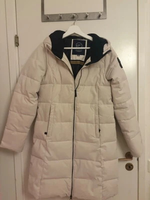 Beige lång pufferjacka Five Seasons - Säljer en beige lång pufferjacka från Five Seasons med huva och svarta detaljer. Jackan har quiltad design, dragkedja framtill och två fickor. Perfekt för kalla dagar och har en stilren look med logotyp på ärmen. Köpte för 1000kr