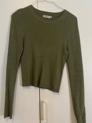 Olivgrön ribbad tröja Stradivarius - Säljer en olivgrön ribbad tröja från Stradivarius i storlek XS. Tröjan har lång ärm, rund hals och är croppad i modellen. Materialet är mjukt och stretchigt, perfekt för att styla med jeans eller kjol. Enkel och snygg design som funkar året runt.