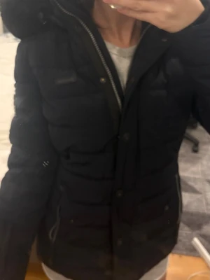 Svart pufferjacka från Navahoo - Säljer en svart pufferjacka från Navahoo med stor fluffig huva i riktig päls. Jackan har dragkedjor och knappar framtill, samt två fickor med dragkedja. Perfekt för kalla dagar och har en snygg look. Köpte för 3000kr.