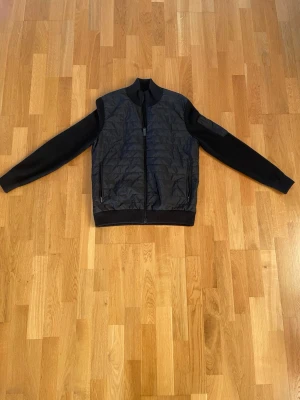 Svart quiltad bomberjacka J.Lindeberg - Svart bomberjacka från J.Lindeberg i storlek M. Framsidan är quiltad med syntetmaterial och har två fickor med dragkedja. Ärmar och rygg är stickade, vilket ger en schysst kontrast. Jackan har ribbad krage och muddar samt JL-logga på ärmen.