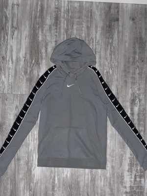 Nike Swoosh Tape Hoodie - Grå Nike hoodie med huva och små nike-loggor på en svart remsa längs armarna. Tröjan är i bra skick, Använd ett par gånger. 