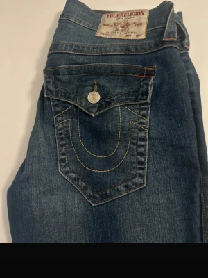 True Religion blå jeans med kontrastsömmar - Säljer ett par klassiska blå jeans från True Religion med ikonisk kontrastsöm på bakfickan och snygga detaljer i gult och orange. Jeansen har normal passform och är tillverkade i slitstarkt denim med knappstängning och fickor fram och bak.