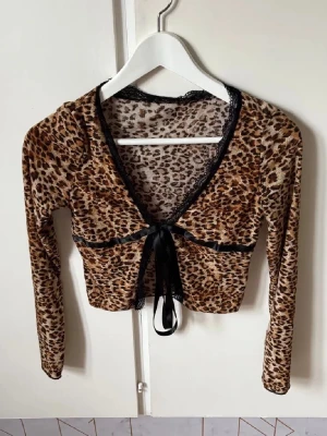 Leopardmönstrad croppad bolerotopp - Säljer en croppad bolerotopp med leopardmönster i bruna och svarta toner. Toppen har lång ärm, v-ringad hals med svart spetskant och en snygg knytning framtill med satinband. Materialet känns mjukt och stretchigt, perfekt för att sticka ut.