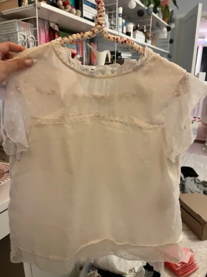 Ljusbeige prickig blus med volang - Säljer en ljusbeige blus med små prickar i tyget och transparenta, korta ärmar. Blusen har en söt volangkant vid halsen och ett tunt, luftigt material som ger en romantisk känsla. Perfekt för dig som gillar detaljer och mjuka färger.