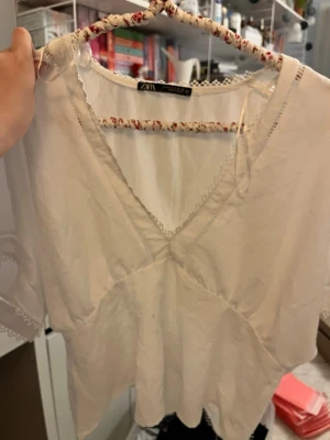 Vit blus med v-ringning från Zara - Säljer en luftig vit blus från Zara med v-ringning och fina spetsdetaljer längs kanterna. Blusen har korta ärmar och är tillverkad i ett lätt bomullstyg, perfekt för varmare dagar. Enkel och stilren design som passar till mycket.