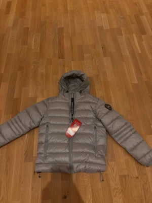 Grå dunjacka från Canada Goose - Säljer en grå dunjacka från Canada Goose med huva och två sidofickor med dragkedja. Jackan har quiltad design, justerbar snörning nedtill och logotyp-patch på ärmen. Perfekt för kalla dagar och har en clean look med diskreta detaljer.