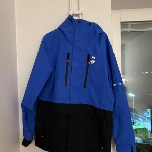 Blå och svart skidjacka i skal från Montec - Snygg tvåfärgad skidjacka i skal från Montec i blått och svart med huva och praktiska dragkedjor framtill. Jackan har lång ärm, justerbara muddar och logga på bröstet och ärmen. Perfekt för dig som gillar att vara ute eller åka skidor och vill ha en cool look.