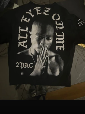 Svart 2Pac långärmad tröja - Svart oversized långärmad tröja med stort 2Pac-tryck och texten 'All Eyez On Me' på ryggen. Framsidan har 2Pac-logga och Tupac Amaru Shakur-signatur i vitt, även signatur på ena ärmen. Snygg streetwear med loose fit och coolt tryck.