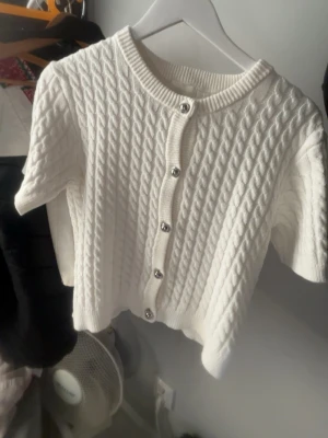 Vit kortärmad kofta med kabelmönster - Säljer en vit, kortärmad kofta med snyggt kabelstickat mönster och silvriga knappar framtill. Tröjan har rund hals och ribbade kanter vid ärmslut och nederkant. Perfekt för dig som gillar en clean och fräsch look.