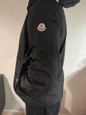 Svart Moncler vindjacka med huva - Snygg svart vindjacka från Moncler med huva och coola vita och röda detaljer på kanten. Jackan har dragkedja, snörning vid huvan och Moncler-logga på ärmen. Perfekt för dig som vill ha en stilren jacka med street vibe.