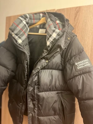 Svart Burberry pufferjacka med rutig krage - Säljer en svart pufferjacka från Burberry med stor patch på ärmen och en snygg rutig krage i grått, vitt och rött. Jackan har dragkedja, knappar och två fickor med dragkedja. Perfekt för kalla dagar och har en riktigt schysst streetstil.