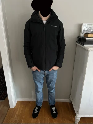 Svart pufferjacka Peak Performance - Svart pufferjacka från Peak Performance med huva och dragkedja framtill. Jackan har en clean look, diskret logga på bröstet och är fodrad för extra värme. Materialet är syntet och polyester, perfekt för kalla dagar. Snygg passform och praktiska fickor.