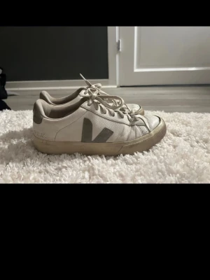 Vita Veja sneakers med gröna detaljer - Säljer ett par vita Veja-sneakers med grön Veja Logga i använt skick. Modellen är klassisk och funkar till allt. Skorna är slitna med smutsfläckar, creases och märken på sulan samt vid tån (se bilder). Innersulan hel, loggan syns tydligt bak. Sulan är använd men fortfarande hel (gått upp lite längst framme vid tårna)   Pris går att diskutera.