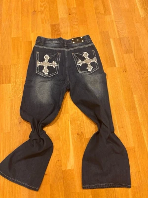Bootcut jeans från Miss Me med dekorerade fickor - Säljer ett par mörkblå bootcut jeans från Miss Me med stora kors i silver och nitar på bakfickorna. Jeansen har vita kontrastsömmar och klassisk femficksdesign. Materialet är jeans och de har dragkedja framtill.