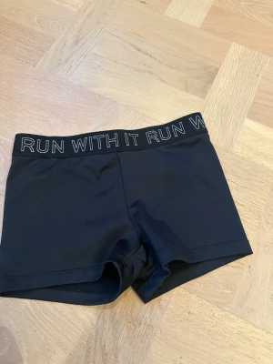 Svarta träningsshorts från Cubus - Snygga svarta träningsshorts från Cubus med elastisk midja där det står 'RUN WITH IT' i vitt. Tillverkade i mjuk polyester och elastan som ger bra stretch och komfort. Perfekta för sport och aktivitet.