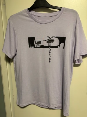 Ljuslila t-shirt med anime-tryck - Säljer en ljuslila t-shirt med korta ärmar och ett svart anime-inspirerat tryck med japansk text på bröstet. T-shirten har rund hals och är gjord i mjuk bomull, perfekt för dig som gillar stilrena och unika motiv.