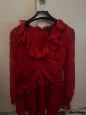 Röd volangklänning från Boohoo - Supersöt röd kortklänning från Boohoo med volanger, djup v-ringning och rynkade detaljer framtill. Klänningen har långa ärmar i transparent material med blommigt burnout-mönster och knappar framtill. Perfekt för dig som vill sticka ut!