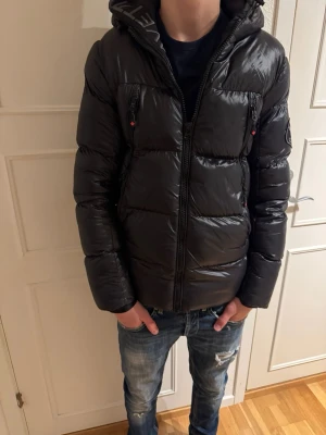 Svart pufferjacka från Zavetti Canada - Säljer en svart pufferjacka från Zavetti Canada med glansig finish och broderad logga på ärmen. Jackan har huva, flera dragkedjeförsedda fickor och en schysst, lite oversized passform. Perfekt för kalla dagar och riktigt snygg till jeans.