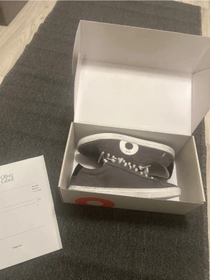 Grå sneakers från Oliver Cabell - Snygga grå sneakers från Oliver Cabell med vit rund detalj på sidan och vita snören. Skorna har en platt vit sula och är tillverkade i mocka med inslag av skinn. Perfekta för dig som gillar stilrena och moderna sneakers med unik design.