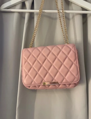 Rosa quiltad axelväska med kedja - Säljer en ljusrosa axelväska med quiltat mönster och guldfärgad kedja som axelrem. Väskan har en rektangulär form, ett innerfack med dragkedja och stängs med ett metallspänne framtill. Materialet är syntet och insidan är fodrad i matchande rosa tyg.