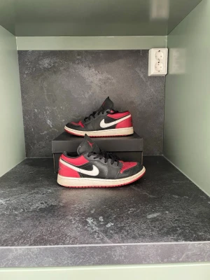 Nike Air Jordan 1 Low svart/röd - Säljer ett par Nike Air Jordan 1 Low sneakers i svart och rött med vit swoosh och sula. Klassisk siluett med snörning och låg profil. Perfekta för dig som gillar streetwear och vill ha en ikonisk sneaker i garderoben.