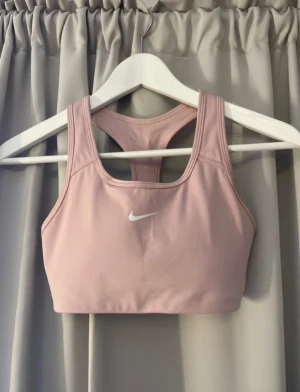 Rosa sport-bh från Nike Dri-Fit - Nike Dri-Fit sport-bh i ljusrosa med klassisk racerback och vit logga framtill. Tillverkad i ett mjukt och stretchigt syntetmaterial som andas och håller dig torr under träningen. Perfekt för gymmet eller löprundan.