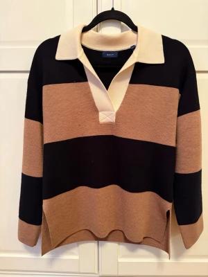 Randig tröja från Gant - Säljer en snygg randig tröja från Gant i beige, svart och brunt. Tröjan har klassisk krage, v-ringning och långa ärmar. 