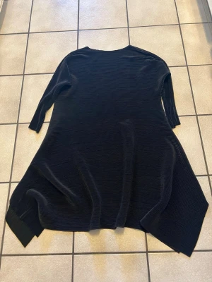 Svart ribbad klänning från YICHU storlek XL/XXL - Svart klänning med ribbad struktur från YES YICHU. Modellen har asymmetrisk nederkant och trekvartsärm. Tillverkad i mjuk bomull med lite stretch från spandex, vilket gör den bekväm och följsam. Perfekt för dig som gillar stilrena och enkla plagg med en twist.