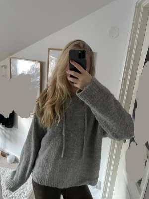 Grå stickad hoodie med huva - Mysig grå stickad hoodie med huva och snörning. Tröjan har en loose passform och långa ärmar, perfekt för kyliga dagar. Materialet är mjukt och fluffigt, vilket gör den extra skön att ha på sig.