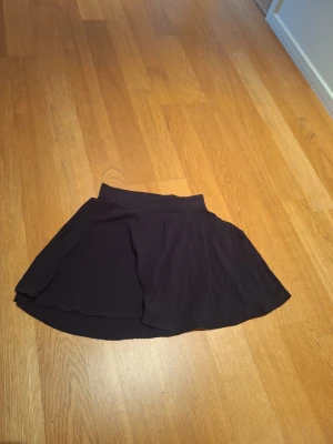 Svart kortkjol från Emoi - Säljer en svart kortkjol från Emoi by Emonite i storlek 164/14. Kjolen har en enkel och klassisk design med lätt utställd form och bred linning. Perfekt att matcha med sneakers eller boots för en chill look.