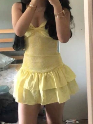 Gul kort volangklänning med knyt - Säljer en söt gul kort klänning med volanger och smala axelband. Klänningen har omlottknytning i ryggen och en lätt ribbad struktur. Perfekt för dig som gillar ljusa färger och vill ha en feminin look med volangdetaljer. Färgen är mer lik den andra bilden!