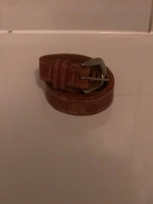 Brun skärp i skinn från Esprit - Säljer ett brunt skärp från Esprit i äkta skinn med kraftig metallspänne och vita dekorativa sömmar längs kanterna. Skärpet är cirka 90 cm långt och har klassisk design som funkar perfekt rill jeans 👌.