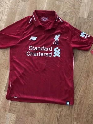 Liverpool tröja Firmino #9 New Balance - Liverpool FC matchtröja i röd färg med Firmino och nummer 9 på ryggen. Tröjan har klassisk krage, vita detaljer och tryckta loggor från New Balance och Standard Chartered. Tillverkad i lätt polyester för bästa komfort. Perfekt för fotbollsfans!