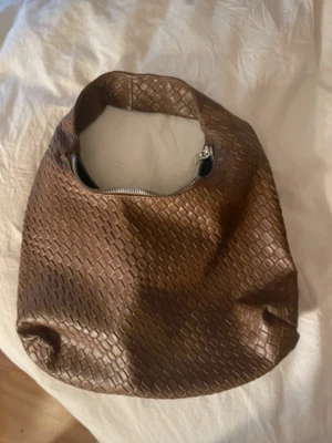 Brun axelväska med flätad struktur - Säljer en brun axelväska med snygg flätad struktur och rundad form. Väskan har en praktisk dragkedja upptill och är tillverkad i skinnimitation. Perfekt storlek för att få plats med allt du behöver och passar till många olika stilar.