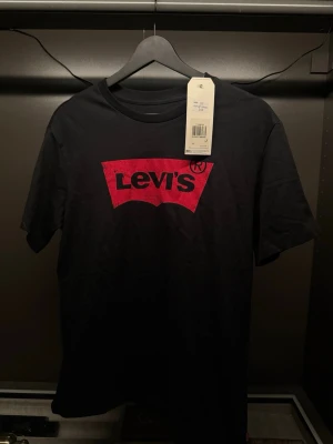 Svart Levi's t-shirt med röd logga - Klassisk svart t-shirt från Levi's i bomull med den ikoniska röda loggan tryckt på bröstet. T-shirten har rund halsringning och korta ärmar, perfekt för en avslappnad och stilren look.