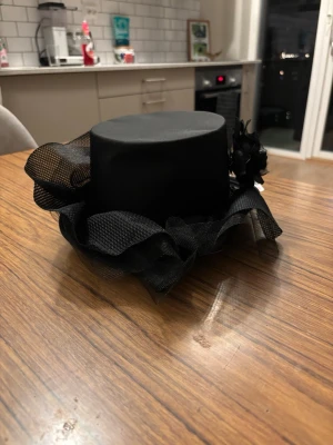 Svart cylinderhatt med tyll och blomma - Säljer en svart cylinderhatt med dekorativt tyll och en stor svart tygblomma på sidan. Hatten har en klassisk form och är perfekt för att sticka ut på temafester eller maskerad. Materialet är syntet och tyll, vilket ger en cool och dramatisk look.