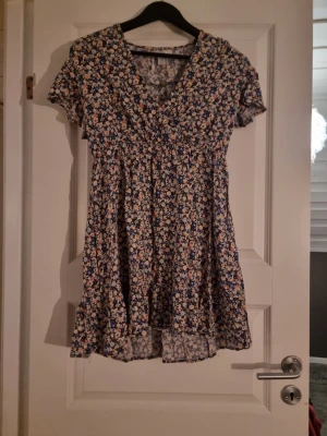 Blommig kortklänning från Amisu - Säljer en söt kortklänning från Amisu med blommigt mönster i blått, beige, orange och vitt. Klänningen har v-ringad hals, korta ärmar och volangdetalj nedtill. Materialet är lätt och känns som polyester, perfekt för varma dagar.