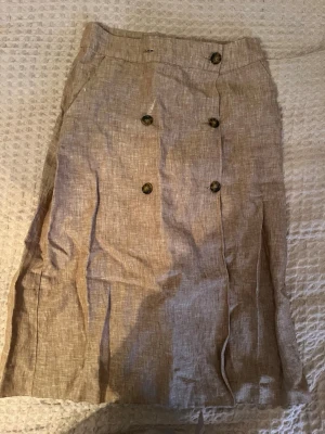 Beige linnekjol från Mango - Säljer en beige kjol (går till knäna ungefär) från Mango i storlek S. Kjolen är tillverkad i linne och har knappar framtill. Perfekt för dig som gillar en enkel och stilren look med naturliga material.