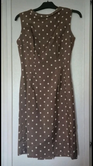 Brun prickig ärmlös klänning - Retroinspirerad brun klänning med vita prickar och ärmlös design. Klänningen har en klassisk, något figurnära passform och går till knäna. Dragkedja bak för enkel påklädning. Klänningen saknar märkningar men skulle gissa att den är en stl XS/S och den är inte stretchig. De skavanker jag har kunnat se är att slitsen behöver lagas och tyget är lite skadat på höger axel (se bilder).