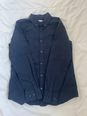 Mörkblå skjorta från 157 - Snygg mörkblå skjorta från 157 med klassisk krage och knappar hela vägen framtill. Skjortan har lång ärm och en bröstficka. Materialet känns som bomull och passformen är normal. Perfekt för dig som gillar stilrena och enkla plagg.