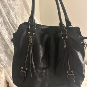 Svart axelväska i skinn med dragkedjor - Snygg svart axelväska i skinn med två långa handtag, dekorativa spännen och dragkedjor framtill. Väskan har även en praktisk ficka med dragkedja på baksidan och coola detaljer i metall. Perfekt för dig som vill ha en stilren och rymlig väska. 